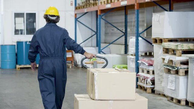 Back view of senior worker pulling trolley with cardboard boxes. Man in helmet preparing goods for transportation. Warehouse, transportation concept.