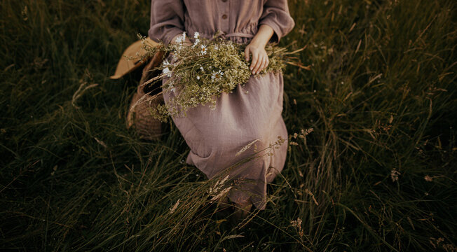 Beautiful woman in linen dress sitting on rustic chair among grass with bouquet of wildflowers in summer meadow. Young female relaxing in countryside. Atmospheric stylish dreamy image