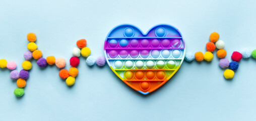 on a blue background lies a toy pop it in the shape of a heart in a rainbow color and a cardiogram made of multi-colored balls. toy addiction concept