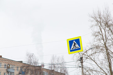 A pedestrian crossing road sign hangs over the carriageway. Against the background of the gray sky. City life and road traffic concept. Elements of a modern metropolis.