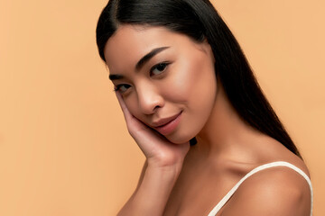 Young Asian woman with clean healthy glowing skin in white top isolated on beige background. Facial...