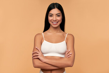 Young Asian woman in white lingerie crossed her arms on a beige background. Sports, healthy lifestyle. Spa body care. Plastic surgery. Cosmetology concept.