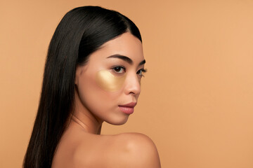 Young Asian woman in white lingerie and clean radiant skin with moisturizing patches under the eyes on a beige background. Spa care, cosmetology.