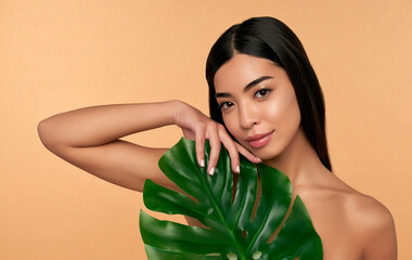 Young Asian woman with clean radiant face skin with green leaf on beige background. Spa care, facial skin care, beauty cosmetology. Natural cosmetics concept.