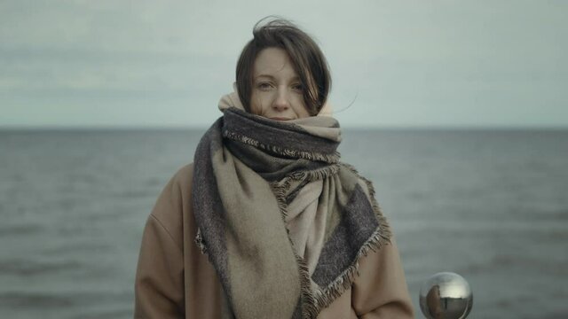 Portrait of woman in warm beige oversize coat and scarf standing against ocean. Close up portrait of young woman with dark hair blowing in wind looking at the camera 