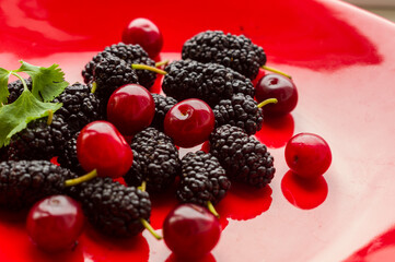 Ripe large black mulberries and red cherries on a red plate, close-up.