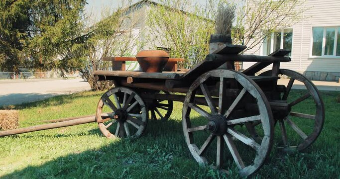 In the yard there is a cart made of wood. It is half-lit by the midday sun. On the cart is a pitcher, a barrel of dead wood and a small bench. Next to it is a rectangular haystack.