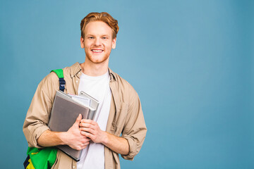 Portrait of happy smiling young student standing with backpack and folders isolated over blue...