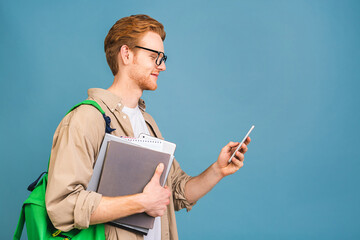 Portrait of happy smiling young student standing with backpack and folders isolated over blue background. Using mobile phone.