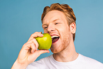 Diet concept and healthy lifestyle. Cheerful beautiful young man eating apple, isolated over blue background.