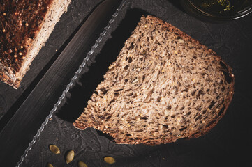 slice of rustic natural yeast-free bread with flax, poppy seeds, sesame seeds, millet, pumpkin and sunflower seeds, with olive oil in a glass jar, and a knife, on a black background, hard light