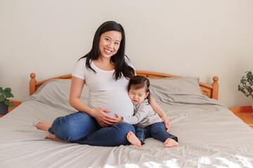 Asian Chinese pregnant woman with toddler girl sitting on bed at home. Girl daughter kid hugging...