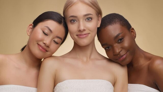 Three young multiethnic women posing wrapped in towels over beige studio background, embracing and smiling at camera