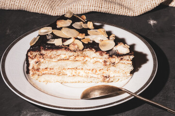Piece of layered homemade sponge cake on a white saucer with a spoon and a linen napkin, black background, hard light, photo in a low key.