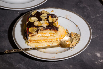 Piece of layered homemade sponge cake on a white saucer with a spoon and a linen napkin, black background, hard light, photo in a low key.