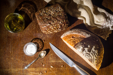 Still life - yeast-free buckwheat bread, black bread with sunflower seeds, olive oil and coarse salt in glass jars, a knife, and a linen napkin on a wooden board, wooden background, hard light