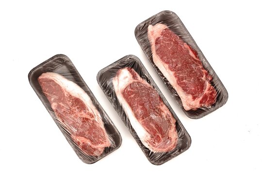 Close-up Striploin Beef Steaks on Foam Tray With Plastic Wrapping Film Isolated on White Background. Commercial Butcher Cut Of Marbled Beef Steaks, Overhead View. Set Of Wrapping Beef Steaks.