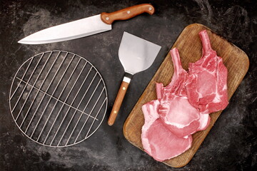 Raw Pork Bone Steaks and Grill Cooking Tools On Wooden Chopping Board. Tomahawk Pork Steaks On Chopping Board, Overhead View. Raw Ribeye Pork Steaks On Wood Board And Black Background, Top View.