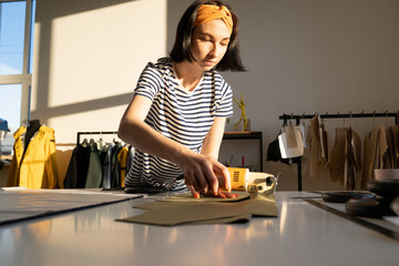 Professional young female tailor making clothing samples in factory or design studio with electric cutter scissors hold paper pattern on table in workshop. Casual designer or seamstress worker at work
