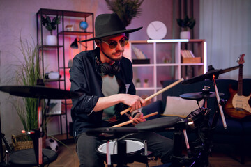 Handsome young man stylish hat and sunglasses using electronic drums for performing new song. Inspired male artist playing drums at dark atmosphere.