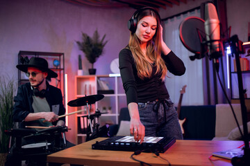 Adorable young woman in headphones working on mixing board while stylish man in hat and sunglasses...