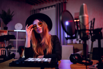 Fototapeta na wymiar Confident music maker in trendy hat and eyeglasses working on dj controller and laptop while creating new track. Charming woman sitting at table and looking at camera.