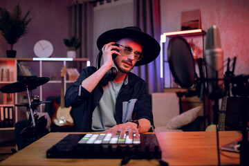Handsome male hipster in black hat and sunglasses creating music using dj controller and...