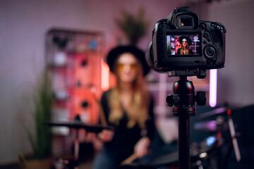 Blur background of female artist in hat and glasses playing electronic drums and recording video. Focus on modern digital camera that fixed on tripod. Online tutorial concept.