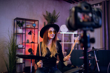 Stylish woman recording music video blog while playing electronic drums at home. Female musician in...