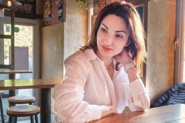 Beautiful young woman with red hair in a white shirt sits at a wooden table in a bar and is thoughtful. Young woman portrait. Woman waiting for a meeting.