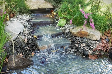 Small Waterfall and Cascade Stream of Water in a Home Private Garden.  Artificial Stream in Summer Backyard Garden. Designed Back Yard Garden with Decorative Pond. PVC Pond Garden With Natural Stones.