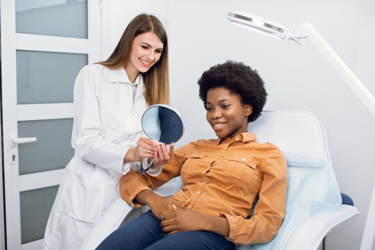 Consultation In Cosmetology Clinic. Female Beauty Doctor Talking With Patient, Young African American Woman, Looking At The Mirror And Satisfied After The Beauty Procedures