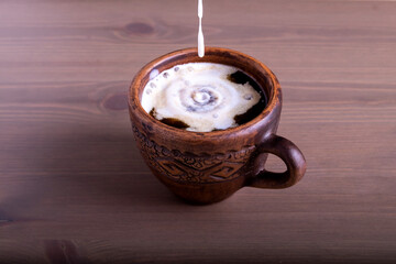 A close-up of hot coffee with milk in a fired earthenware mug. A trickle of milk or cream is poured into the coffee. Natural wood background.