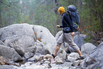 Young man with backpack and in hiking clothes carefully crosses the river in the mountains. Stylish traveler doing trekking along the trail in the national park, it is surrounded by rocks and forests.