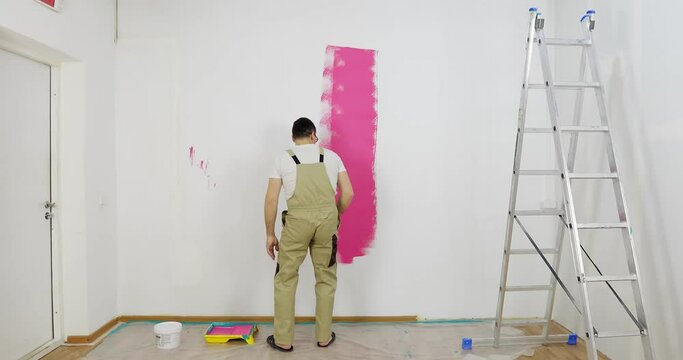 Painter man painting the wall in home, with paint roller and pink color paint. Room renovations at house. Offering professional painting services.