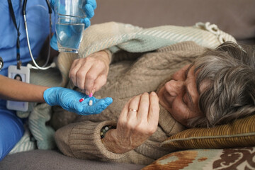 A young nurse is caring for an elderly  80-year-old woman at home. She holds a glass of water and gives medicine pills a pensioner  retired woman who lies and rests in bed.