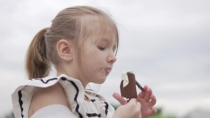 Funny little girl eats ice cream on a stick in the summer on the street.