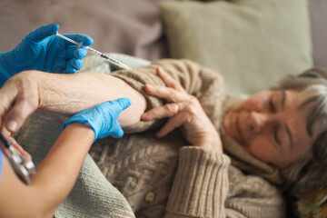 The nurse makes an injection with the vaccine to the patient at home. A young doctor in a blue suit holds the hand of an elderly  woman and makes injection with a syringe. Medicine and healthcare.