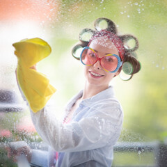 Young woman with curlers in her hair cleans a window with rag and wiper in hand