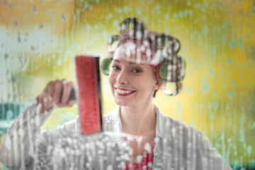 Young woman with curlers in her hair cleans a window with rag and wiper in hand