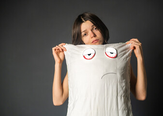 Young girl with a disgruntled face holds a pillow, on which a displeased face and tired eyes are...