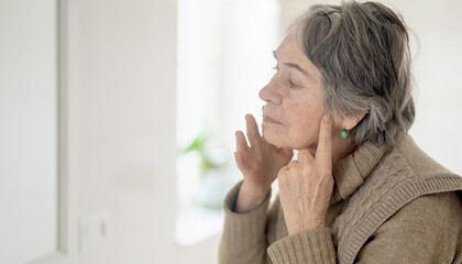 An elderly woman puts her fingers on the lymph nodes in her neck and looks at herself in the mirror. The pensioner takes care, monitors thyroid hormones and the health of the endocrine system.