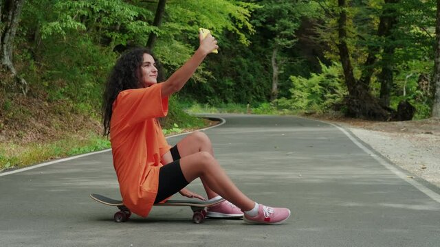 young woman skater is taking selfies sitting on skateboard in park, female skateboarder