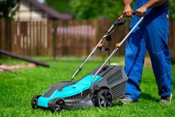 Close-up of a man in overalls with a lawn mower cutting green grass in a modern garden. Lawn mowing machine.