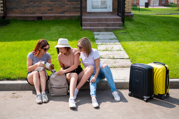 Three Caucasian women and a dog go on a trip. The girls are sitting on the curb with suitcases and waiting for a taxi. Summer vacation concept together with girlfriends