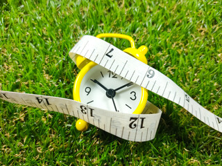 Alarm clock and measuring tape on grass background. Time and healthcare concept. Yellow alarm clock...