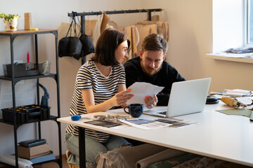 Young tailors work in atelier with laptop, fabric swatches on clothes collection creation. Team of...