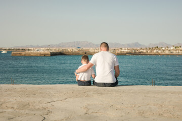 Father and  son  spending time together sea vacation. Little boy and Dad sitting on beach and looks out to sea. Back view. Family with one child. Happy childhood with daddy.