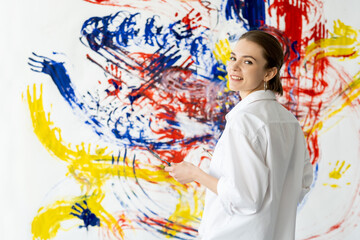 Obraz na płótnie Canvas Hand painting. Art therapy. Relaxing leisure. Happy smiling artist woman in white shirt on colorful yellow blue red abstract artwork stained wall background.