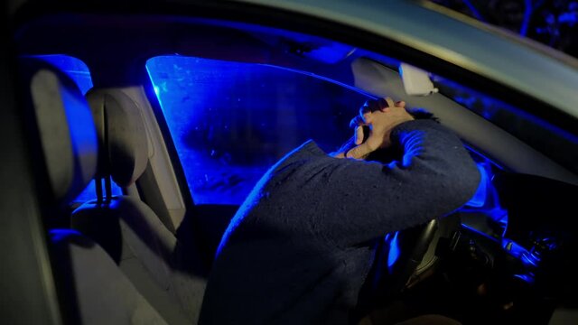 Drunk driver in black sweater inside car salon looks at flashing police lights and puts crossed hands behind head at night closeup